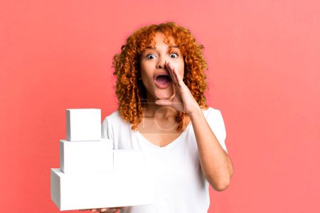Photo for Red hair pretty woman feeling happy,giving a big shout out with hands next to mouth. blank packages boxes concept - Royalty Free Image