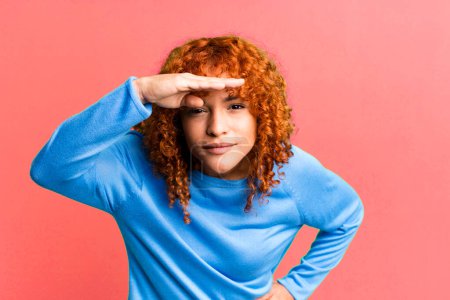Photo for Redhair pretty woman looking bewildered and astonished, with hand over forehead looking far away, watching or searching - Royalty Free Image