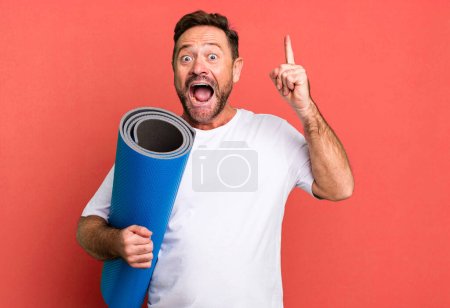 Téléchargez les photos : Homme d'âge moyen se sentant comme un génie heureux et excité après avoir réalisé une idée. avec un tapis de yoga. concept de fitness - en image libre de droit