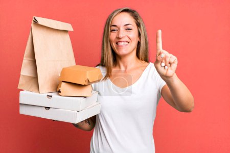 Foto de Bonita mujer rubia sonriendo y buscando amigable, mostrando el número uno. entrega comida para llevar concepto de paquetes - Imagen libre de derechos
