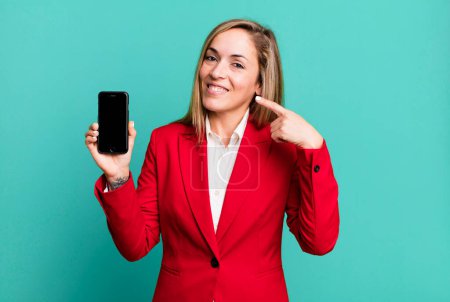 Photo for Pretty blonde woman smiling confidently pointing to own broad smile. businesswoman and a phone concept - Royalty Free Image