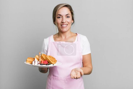 Photo for Pretty blonde woman smiling happily with friendly and  offering and showing a concept. home made waffles concept - Royalty Free Image