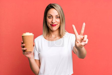 Foto de Bonita mujer rubia sonriendo y buscando amigable, mostrando el número dos con un café caliente - Imagen libre de derechos
