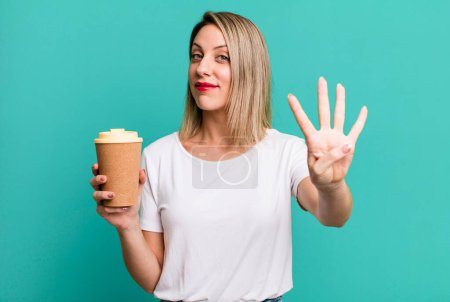 Foto de Bonita mujer rubia sonriendo y buscando amigable, mostrando el número cuatro con un café caliente - Imagen libre de derechos