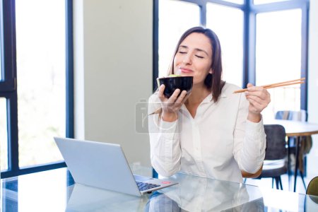 Foto de Pretty young adult woman teleconmmuting with a laptop and eating japanese noodles - Imagen libre de derechos