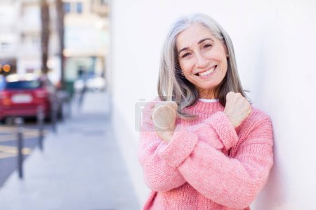 Téléchargez les photos : Senior retraité jolie femme aux cheveux blancs souriant joyeusement et célébrant, avec les poings serrés et les bras croisés, se sentant heureux et positif - en image libre de droit