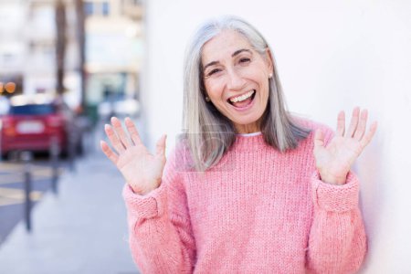 Photo for Senior retired pretty white hair woman looking happy and excited, shocked with an unexpected surprise with both hands open next to face - Royalty Free Image