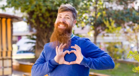 Téléchargez les photos : Cheveux roux homme barbu souriant et se sentant heureux, mignon, romantique et amoureux, faisant forme de coeur avec les deux mains - en image libre de droit