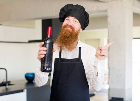Foto de Hombre de pelo rojo sonriendo alegremente, sintiéndose feliz y señalando hacia un lado con una botella de vino. concepto de chef - Imagen libre de derechos