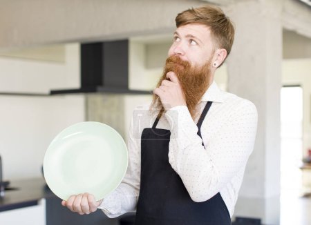 Foto de Hombre de pelo rojo sonriendo con una expresión feliz y segura con la mano en la barbilla con un plato vacío. concepto de chef - Imagen libre de derechos