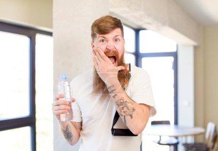 Foto de Hombre de pelo rojo con la boca y los ojos bien abiertos y la mano en la barbilla con una botella de agua. concepto de fitness - Imagen libre de derechos