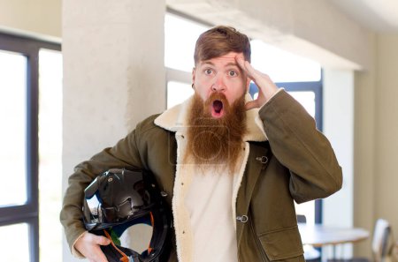 Téléchargez les photos : Homme aux cheveux roux regardant heureux, étonné et surpris avec un casque de moto - en image libre de droit