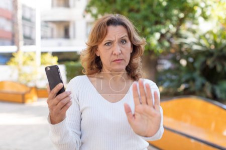 Téléchargez les photos : Moyen âge jolie femme regardant sérieux montrant la paume ouverte faisant geste d'arrêt. en utilisant son smartphone - en image libre de droit