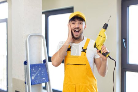 Photo for Young handsome man feeling happy and astonished at something unbelievable. handyman and drill concept - Royalty Free Image