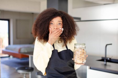 Téléchargez les photos : Jolie afro femme noire couvrant la bouche avec une main et une expression choquée ou surprise. concept de cookies maison - en image libre de droit