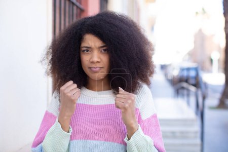 Téléchargez les photos : Afro-américaine jolie femme à l'air confiante, en colère, forte et agressive, avec les poings prêts à se battre en position de boxe - en image libre de droit