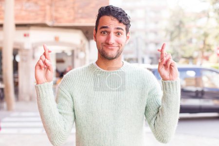 Téléchargez les photos : Jeune homme hispanique croisant les doigts anxieusement et espérant bonne chance avec un regard inquiet - en image libre de droit