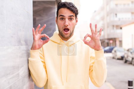 Photo for Young hispanic man feeling shocked, amazed and surprised, showing approval making okay sign with both hands - Royalty Free Image