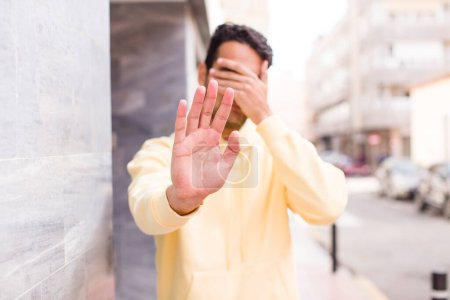 Foto de Joven hispano cubriendo la cara con la mano y poniendo otra mano delante para detener la cámara, negando fotos o imágenes - Imagen libre de derechos
