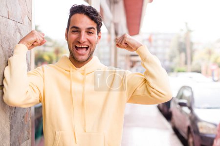 Téléchargez les photos : Jeune homme hispanique se sentant heureux, satisfait et puissant, flexion ajustement et biceps musculaires, regardant fort après la salle de gym - en image libre de droit