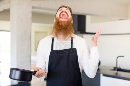 Photo for Red hair man screaming with hands up in the air in a kitchen. chef concept - Royalty Free Image