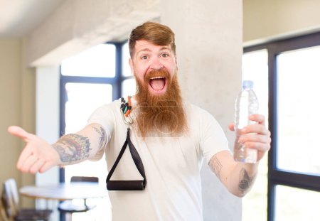 Foto de Hombre pelirrojo sonriendo felizmente y ofreciendo o mostrando un concepto con una botella de agua. concepto de fitness - Imagen libre de derechos
