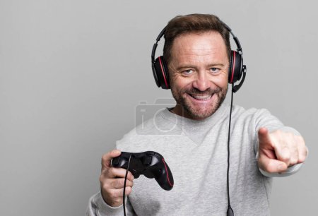 Foto de Hombre de mediana edad apuntando a la cámara eligiéndote. concepto de jugador con un control y auriculares - Imagen libre de derechos