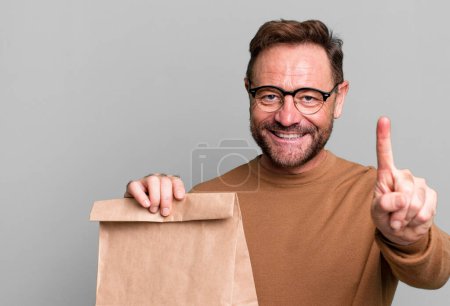 Foto de Hombre de mediana edad sonriendo orgullosamente y con confianza haciendo número uno. llevar bolsa de papel - Imagen libre de derechos