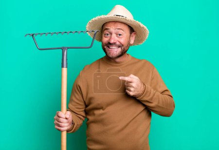 Foto de Hombre de mediana edad sonriendo alegremente, sintiéndose feliz y señalando hacia un lado. granjero con un rastrillo - Imagen libre de derechos