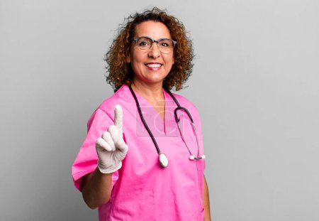 Foto de Bastante mujer de mediana edad sonriendo y buscando amigable, mostrando el número uno. concepto de enfermera - Imagen libre de derechos