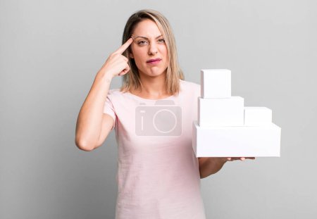 Photo for Pretty blonde woman feeling confused and puzzled, showing you are insane with a set of blank packagings - Royalty Free Image