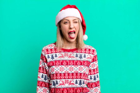 Téléchargez les photos : Jolie femme blonde avec une attitude joyeuse et rebelle, plaisanter et sortir la langue. Noël et santa chapeau concept - en image libre de droit