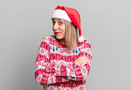 Foto de Bonita mujer rubia encogiéndose de hombros, sintiéndose confundida e incierta. concepto de Navidad y sombrero de santa - Imagen libre de derechos