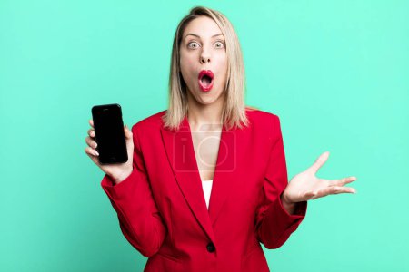 Photo for Pretty blonde woman amazed, shocked and astonished with an unbelievable surprise. businesswoman showing a phone screen - Royalty Free Image