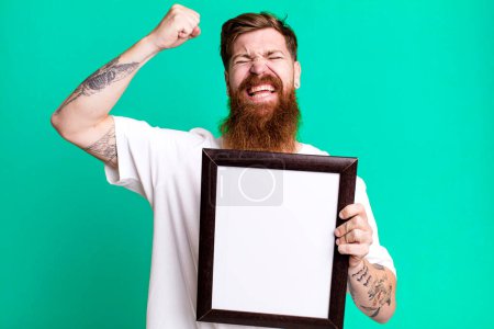 Téléchargez les photos : Jeune adulte cheveux roux barbu cool homme tenant un cadre photo vide - en image libre de droit