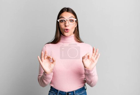 Foto de Pretty young adult woman feeling shocked, amazed and surprised, showing approval making okay sign with both hands - Imagen libre de derechos