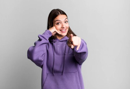 Photo for Pretty young adult woman smiling cheerfully and pointing to camera while making a call you later gesture, talking on phone - Royalty Free Image