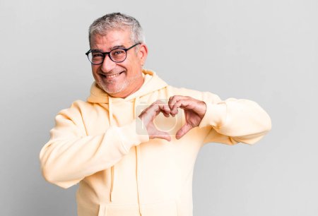 Téléchargez les photos : Homme âgé d'âge moyen souriant et se sentant heureux, mignon, romantique et amoureux, faisant forme de coeur avec les deux mains - en image libre de droit