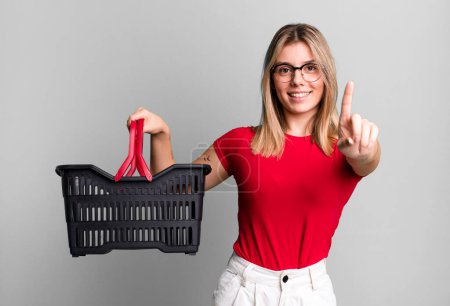 Foto de Young pretty woman smiling and looking friendly, showing number one. empty shopping basket concept - Imagen libre de derechos