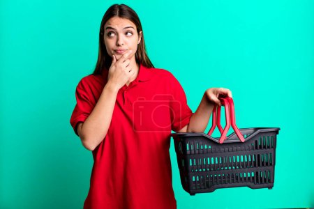 Foto de Young pretty woman thinking, feeling doubtful and confused. empty shopping basket concept - Imagen libre de derechos