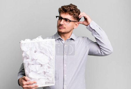 Téléchargez les photos : Jeune homme caucasien adulte souriant joyeusement et rêvant ou doutant avec un concept de poubelle à billes de papier - en image libre de droit