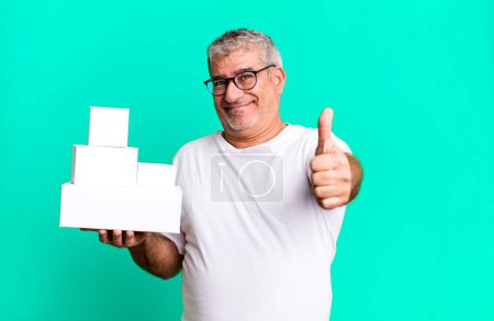 Foto de Hombre mayor de mediana edad sintiéndose orgulloso, sonriendo positivamente con los pulgares hacia arriba. cajas en blanco de diferentes paquetes - Imagen libre de derechos