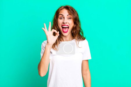 Photo for Young pretty woman feeling successful and satisfied, smiling with mouth wide open, making okay sign with hand - Royalty Free Image