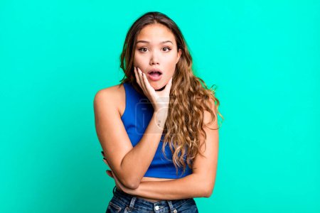 Photo for Young pretty woman open-mouthed in shock and disbelief, with hand on cheek and arm crossed, feeling stupefied and amazed - Royalty Free Image