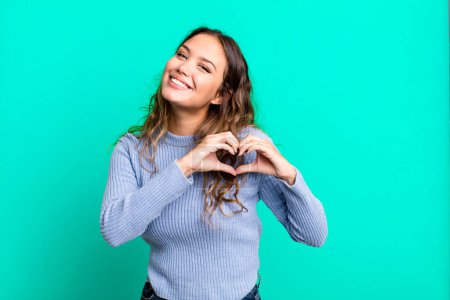 Photo for Young pretty woman smiling and feeling happy, cute, romantic and in love, making heart shape with both hands - Royalty Free Image