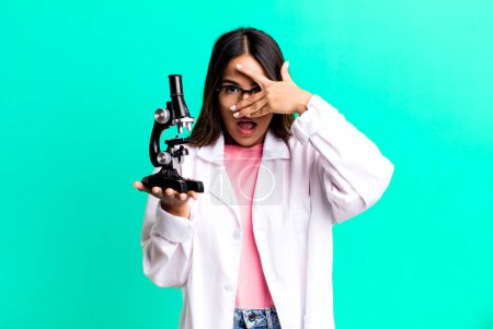 Photo for Hispanic pretty woman looking shocked, scared or terrified, covering face with hand. scients student with a microscope - Royalty Free Image