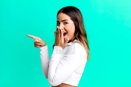 Photo for Hispanic pretty woman feeling happy, shocked and surprised, covering mouth with hand and pointing to lateral copy space - Royalty Free Image