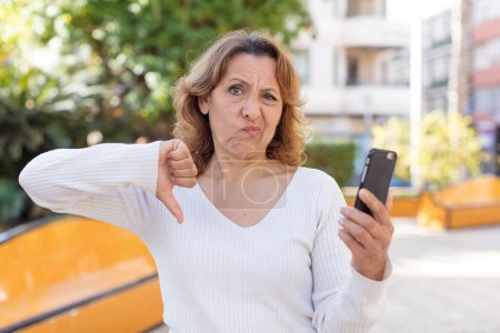 Photo for Middle age pretty woman feeling cross,showing thumbs down. using her smartphone - Royalty Free Image