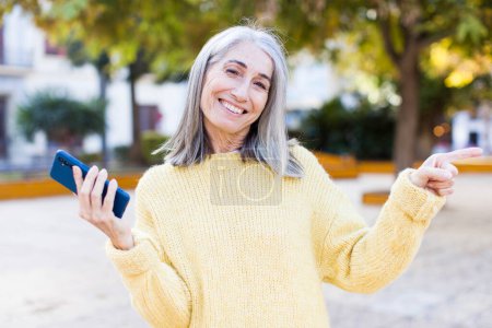 Foto de Mujer bastante mayor sonriendo alegremente, sintiéndose feliz y señalando a un lado con un teléfono inteligente - Imagen libre de derechos