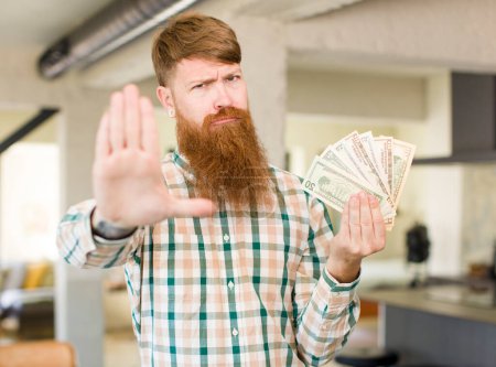 Téléchargez les photos : Homme aux cheveux roux regardant sérieusement montrant la paume ouverte faire un geste d'arrêt avec des billets en dollars - en image libre de droit